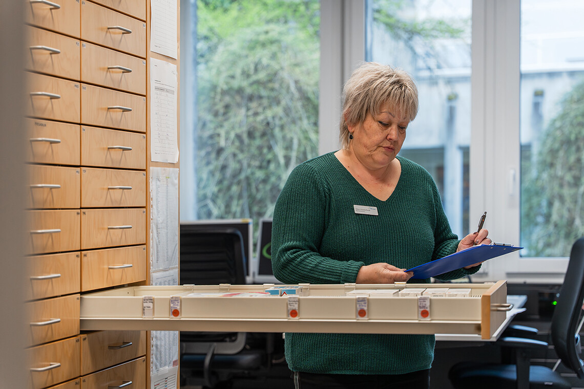 Frau mit grauem Haar im grünen Pullover trägt Namensschild und füllt Formulare in einem Büro, neben ihr ein Holzkästchen mit Beschriftungsboxen und Regalschränke, Fenster mit grünem Blick.