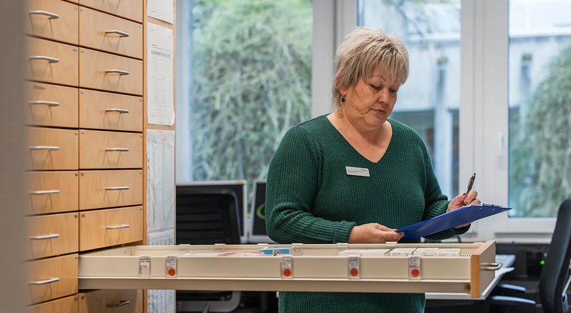 Frau mit grauem Haar im grünen Pullover trägt Namensschild und füllt Formulare in einem Büro, neben ihr ein Holzkästchen mit Beschriftungsboxen und Regalschränke, Fenster mit grünem Blick.