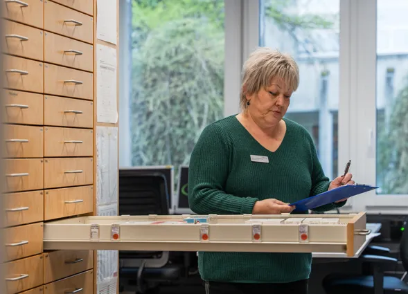 Frau mit grauem Haar im grünen Pullover trägt Namensschild und füllt Formulare in einem Büro, neben ihr ein Holzkästchen mit Beschriftungsboxen und Regalschränke, Fenster mit grünem Blick.