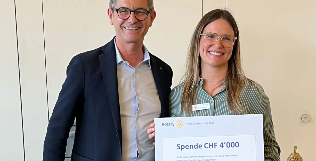 Mann und Frau stehen nebeneinander und halten ein großes Schild mit der Aufschrift „Spende CHF 4'000“ in den Händen. Der Mann trägt einen dunklen Anzug, die Frau ein gestreiftes Hemd. Im Hintergrund sind helle Wände und ein Tisch mit einem kleinen Gong...