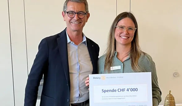 Mann und Frau stehen nebeneinander und halten ein großes Schild mit der Aufschrift „Spende CHF 4'000“ in den Händen. Der Mann trägt einen dunklen Anzug, die Frau ein gestreiftes Hemd. Im Hintergrund sind helle Wände und ein Tisch mit einem kleinen Gong...