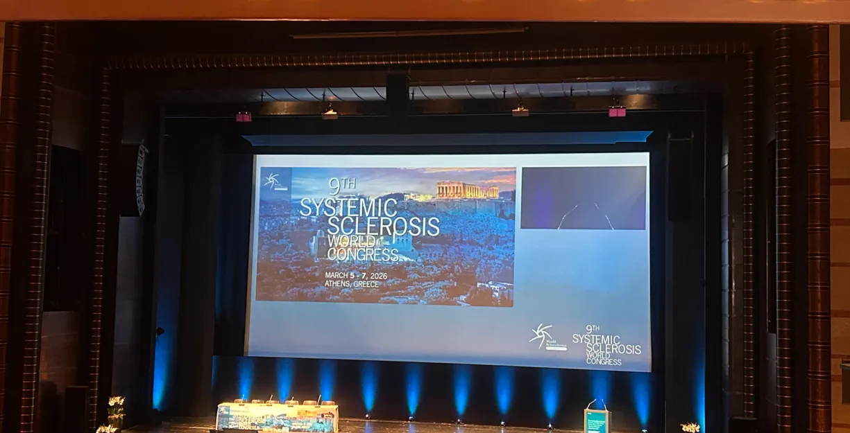 Bühne mit Präsentationsleinwand für den 9. Weltkongress über systemische Sklerose, geplant für den 5. bis 7. März 2026 in Athen, Griechenland. Im Vordergrund ein Rednerpult und dekorative Blumenarrangements.