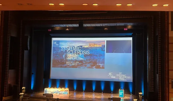Bühne mit Präsentationsleinwand für den 9. Weltkongress über systemische Sklerose, geplant für den 5. bis 7. März 2026 in Athen, Griechenland. Im Vordergrund ein Rednerpult und dekorative Blumenarrangements.