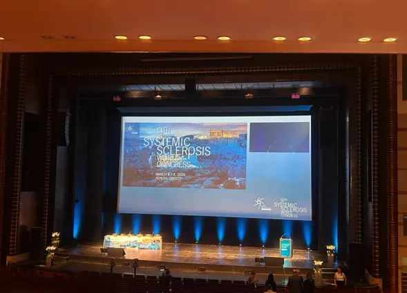 Bühne mit Präsentationsleinwand für den 9. Weltkongress über systemische Sklerose, geplant für den 5. bis 7. März 2026 in Athen, Griechenland. Im Vordergrund ein Rednerpult und dekorative Blumenarrangements.