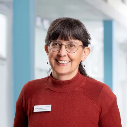 Frau mit kurzen, dunklen Haaren und Brille, trägt einen roten Pullover. Sie lächelt und steht vor einem hellen, modernen Hintergrund. Ein Namensschild ist sichtbar.