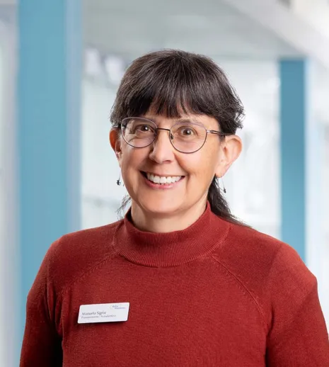 Frau mit kurzen, dunklen Haaren und Brille, trägt einen roten Pullover. Sie lächelt und steht vor einem hellen, modernen Hintergrund. Ein Namensschild ist sichtbar.