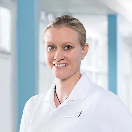 Porträt einer lächelnden Frau mit zusammengebundenem Haar im weißen Kittel vor hellem, modernem Hintergrund.
