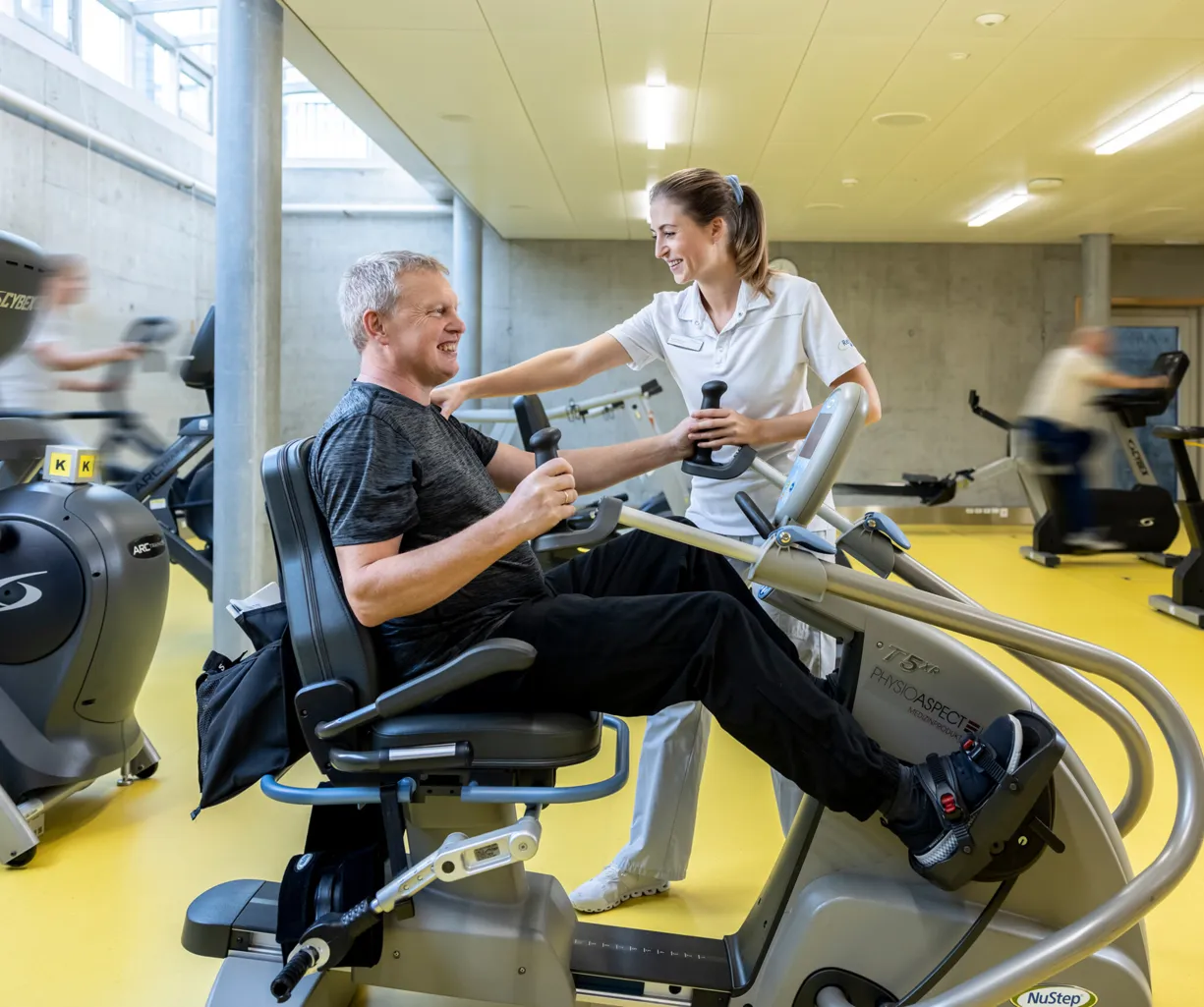 Mann auf einem Trainingsgerät in einem Fitnessstudio, lächelnd und mit Gewichten in den Händen, während eine Trainerin ihm Unterstützung bietet. Im Hintergrund sind weitere Fitnessgeräte und Personen in Bewegung zu sehen. Der Boden ist gelb, und die Wä...