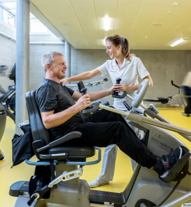 Mann auf einem Trainingsgerät in einem Fitnessstudio, lächelnd und mit Gewichten in den Händen, während eine Trainerin ihm Unterstützung bietet. Im Hintergrund sind weitere Fitnessgeräte und Personen in Bewegung zu sehen. Der Boden ist gelb, und die Wä...