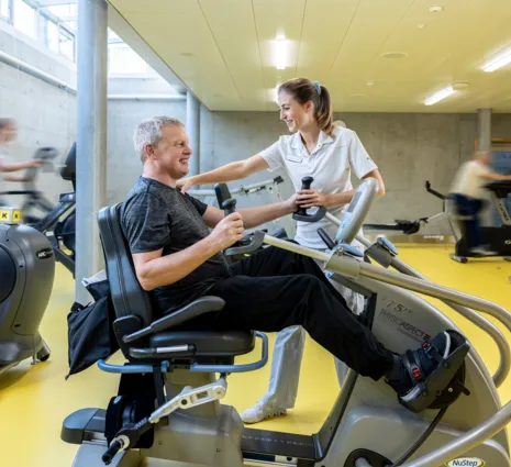 Mann auf einem Trainingsgerät in einem Fitnessstudio, lächelnd und mit Gewichten in den Händen, während eine Trainerin ihm Unterstützung bietet. Im Hintergrund sind weitere Fitnessgeräte und Personen in Bewegung zu sehen. Der Boden ist gelb, und die Wä...