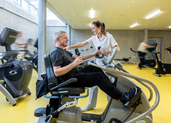 Mann auf einem Trainingsgerät in einem Fitnessstudio, lächelnd und mit Gewichten in den Händen, während eine Trainerin ihm Unterstützung bietet. Im Hintergrund sind weitere Fitnessgeräte und Personen in Bewegung zu sehen. Der Boden ist gelb, und die Wä...