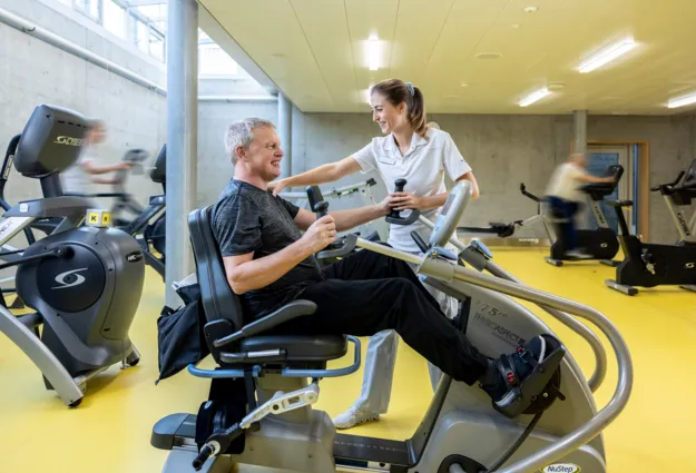 Mann auf einem Trainingsgerät in einem Fitnessstudio, lächelnd und mit Gewichten in den Händen, während eine Trainerin ihm Unterstützung bietet. Im Hintergrund sind weitere Fitnessgeräte und Personen in Bewegung zu sehen. Der Boden ist gelb, und die Wä...