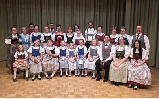 Gruppenfoto von etwa 20 Personen in traditionellen Trachten vor beigem Bühnenvorhang, mehrere mit Schürzen und Dirndl, einige mit Hüten und Wappenplaketten.