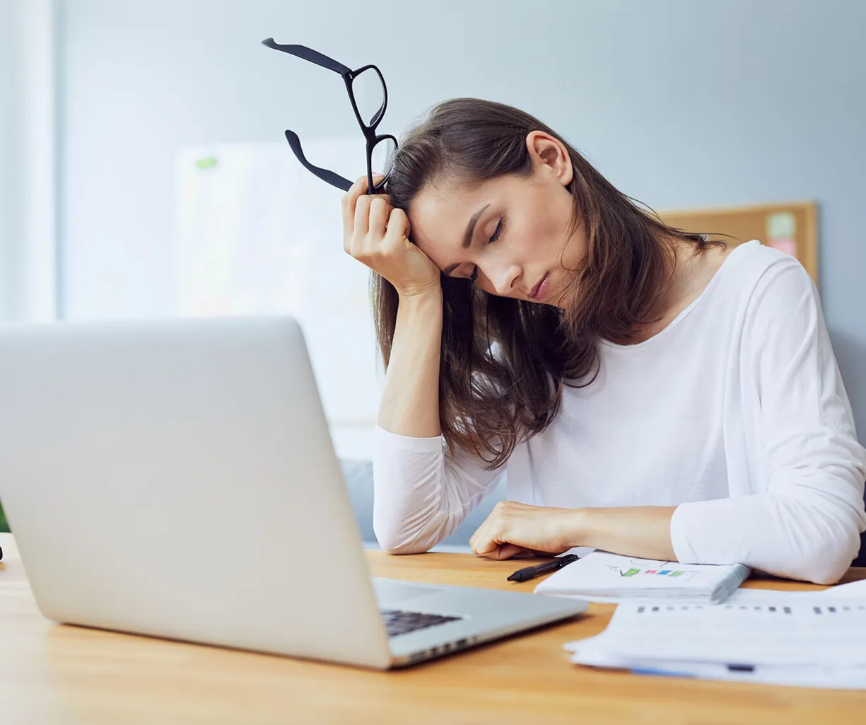 Frau mit langen Haaren sitzt an einem Tisch, hält eine Brille in der Hand und stützt ihren Kopf auf die andere Hand. Vor ihr steht ein Laptop, daneben liegen Unterlagen und ein Taschenrechner. Der Raum ist hell und modern eingerichtet.