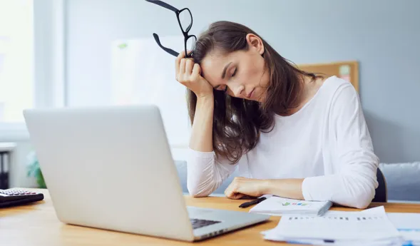 Frau mit langen Haaren sitzt an einem Tisch, hält eine Brille in der Hand und stützt ihren Kopf auf die andere Hand. Vor ihr steht ein Laptop, daneben liegen Unterlagen und ein Taschenrechner. Der Raum ist hell und modern eingerichtet.