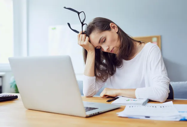 Frau mit langen Haaren sitzt an einem Tisch, hält eine Brille in der Hand und stützt ihren Kopf auf die andere Hand. Vor ihr steht ein Laptop, daneben liegen Unterlagen und ein Taschenrechner. Der Raum ist hell und modern eingerichtet.