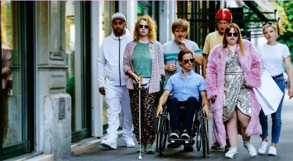 Gruppe gut gekleideter Menschen in der Straße, einige mit roten Kopfbedeckungen und Sonnenbrillen, eine Person im Rollstuhl, bunte Outfits und Mode-Setting vor Gebäuden.