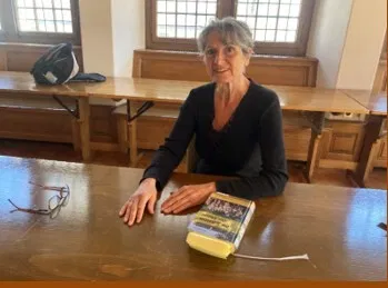 Ältere Frau mit grauem Haar sitzt lächelnd an einem Holztisch; vor ihr liegen eine Brille, ein gelbes Buch und ein weißes Ladekabel. Im Hintergrund: Fenster und Holzmöbel.