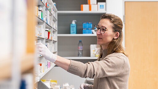 Frau im Labor stellt Regalmaterialien in einem Medikamenten- bzw. Chemie-Lagerraum ein, Hände an einem offenen Fach, im Hintergrund Flaschen, Boxen und Etiketten.