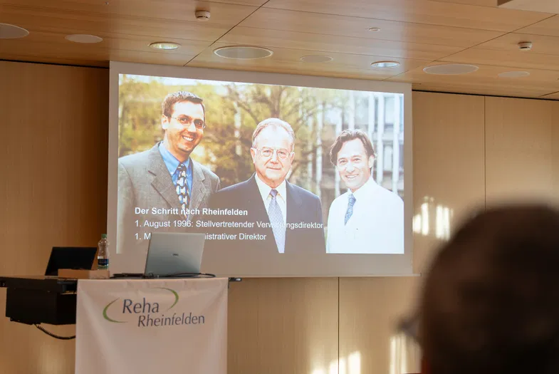 Projektionsleinwand mit Foto dreier Männer und Text zur beruflichen Laufbahn; davor Rednerpult mit Logo „Reha Rheinfelden“ in einem Konferenzraum.