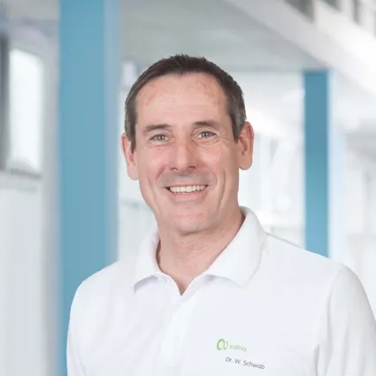 Lächelnder Mann mit kurzen, grauen Haaren, trägt ein weißes Polo-Shirt mit grünem Logo und Namen. Hintergrund mit hellen Wänden und blauen Akzenten.