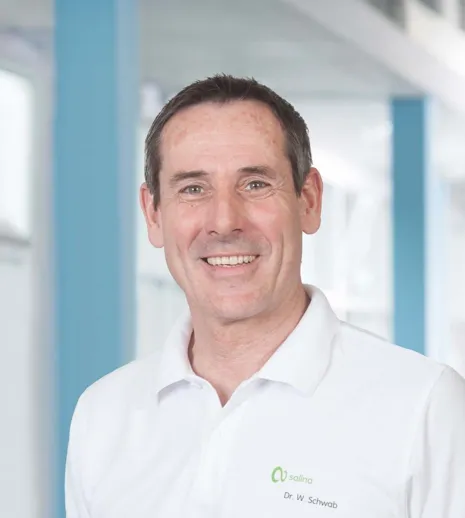 Lächelnder Mann mit kurzen, grauen Haaren, trägt ein weißes Polo-Shirt mit grünem Logo und Namen. Hintergrund mit hellen Wänden und blauen Akzenten.