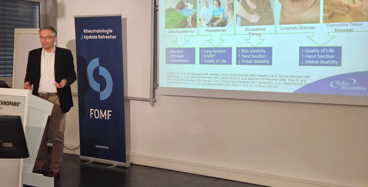 Mann in einem schwarzen Blazer präsentiert vor einer Leinwand mit Informationen zu einem Therapieprogramm für Sklerodermie. Im Hintergrund steht ein Banner mit dem Logo von FOMF. Auf der Leinwand sind verschiedene Therapieansätze und deren Auswirkungen...