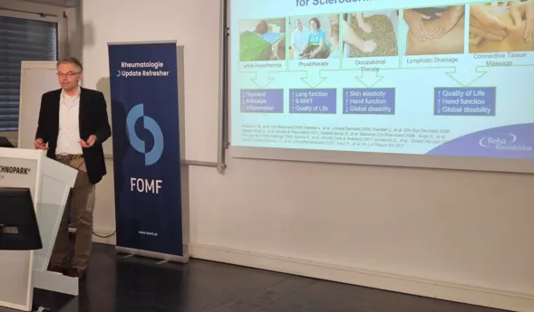 Mann in einem schwarzen Blazer präsentiert vor einer Leinwand mit Informationen zu einem Therapieprogramm für Sklerodermie. Im Hintergrund steht ein Banner mit dem Logo von FOMF. Auf der Leinwand sind verschiedene Therapieansätze und deren Auswirkungen...