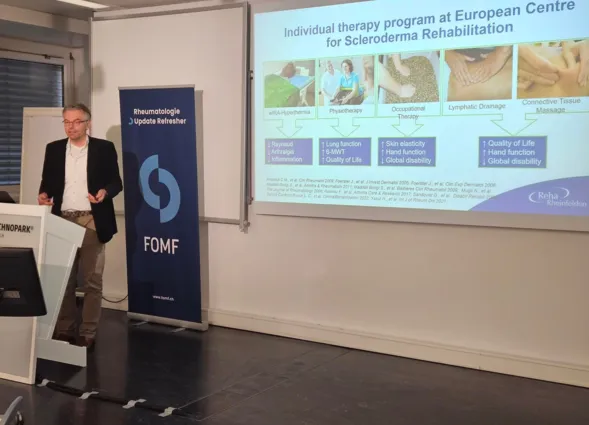 Mann in einem schwarzen Blazer präsentiert vor einer Leinwand mit Informationen zu einem Therapieprogramm für Sklerodermie. Im Hintergrund steht ein Banner mit dem Logo von FOMF. Auf der Leinwand sind verschiedene Therapieansätze und deren Auswirkungen...