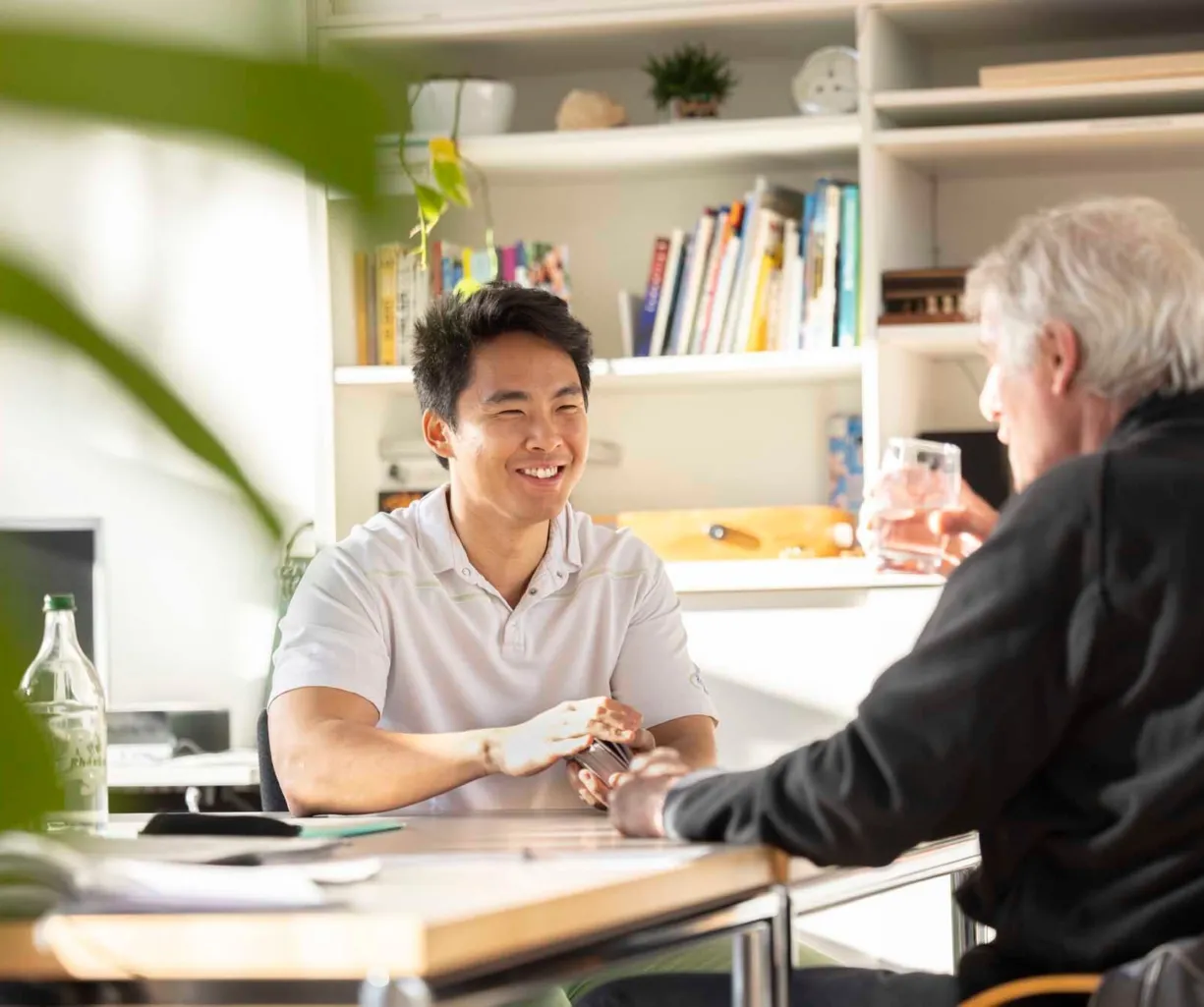 Zwei Männer sitzen an einem Tisch in einem hellen Raum. Der jüngere Mann trägt ein helles Polo-Shirt und lächelt, während er mit dem älteren Mann spricht, der ein schwarzes Oberteil trägt und ein Glas in der Hand hält. Im Hintergrund sind Bücherregale...