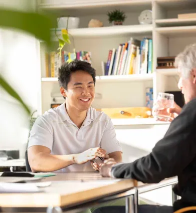 Zwei Männer sitzen an einem Tisch in einem hellen Raum. Der jüngere Mann trägt ein helles Polo-Shirt und lächelt, während er mit dem älteren Mann spricht, der ein schwarzes Oberteil trägt und ein Glas in der Hand hält. Im Hintergrund sind Bücherregale...