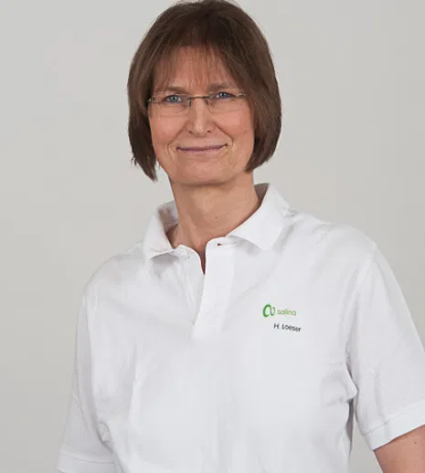 Porträt einer lächelnden Frau mit Brille und kurzem braunem Haar in weißem Poloshirt mit Logo vor grauem Hintergrund.