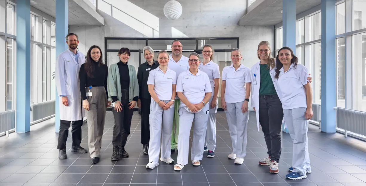 Gruppe von 11 Personen in einem modernen, hellen Raum mit großen Fenstern. Die Personen tragen überwiegend weiße Arbeitskleidung, einige in zivil. Im Hintergrund sind graue Wände und ein minimalistisches Design sichtbar.