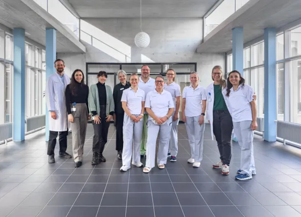 Gruppe von 11 Personen in einem modernen, hellen Raum mit großen Fenstern. Die Personen tragen überwiegend weiße Arbeitskleidung, einige in zivil. Im Hintergrund sind graue Wände und ein minimalistisches Design sichtbar.