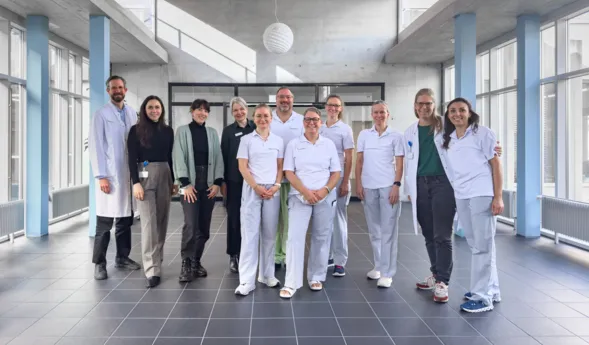 Gruppe von 11 Personen in einem modernen, hellen Raum mit großen Fenstern. Die Personen tragen überwiegend weiße Arbeitskleidung, einige in zivil. Im Hintergrund sind graue Wände und ein minimalistisches Design sichtbar.