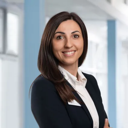 Porträt einer lächelnden Frau mit dunkelbraunem Schulter-Haar im Business-Outfit und weißer Bluse vor hellem, modernem Bürohintergrund.