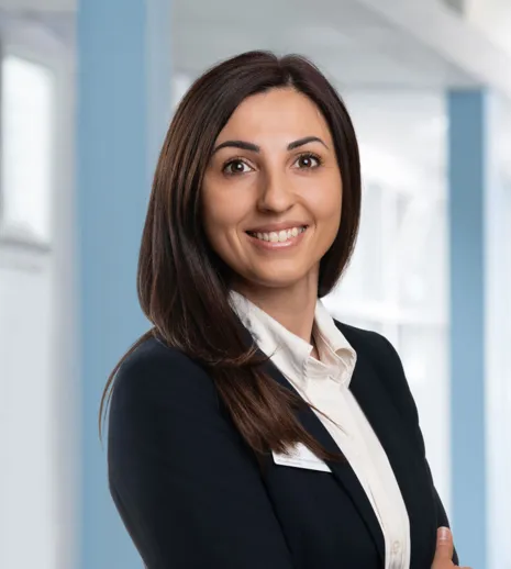 Porträt einer lächelnden Frau mit dunkelbraunem Schulter-Haar im Business-Outfit und weißer Bluse vor hellem, modernem Bürohintergrund.