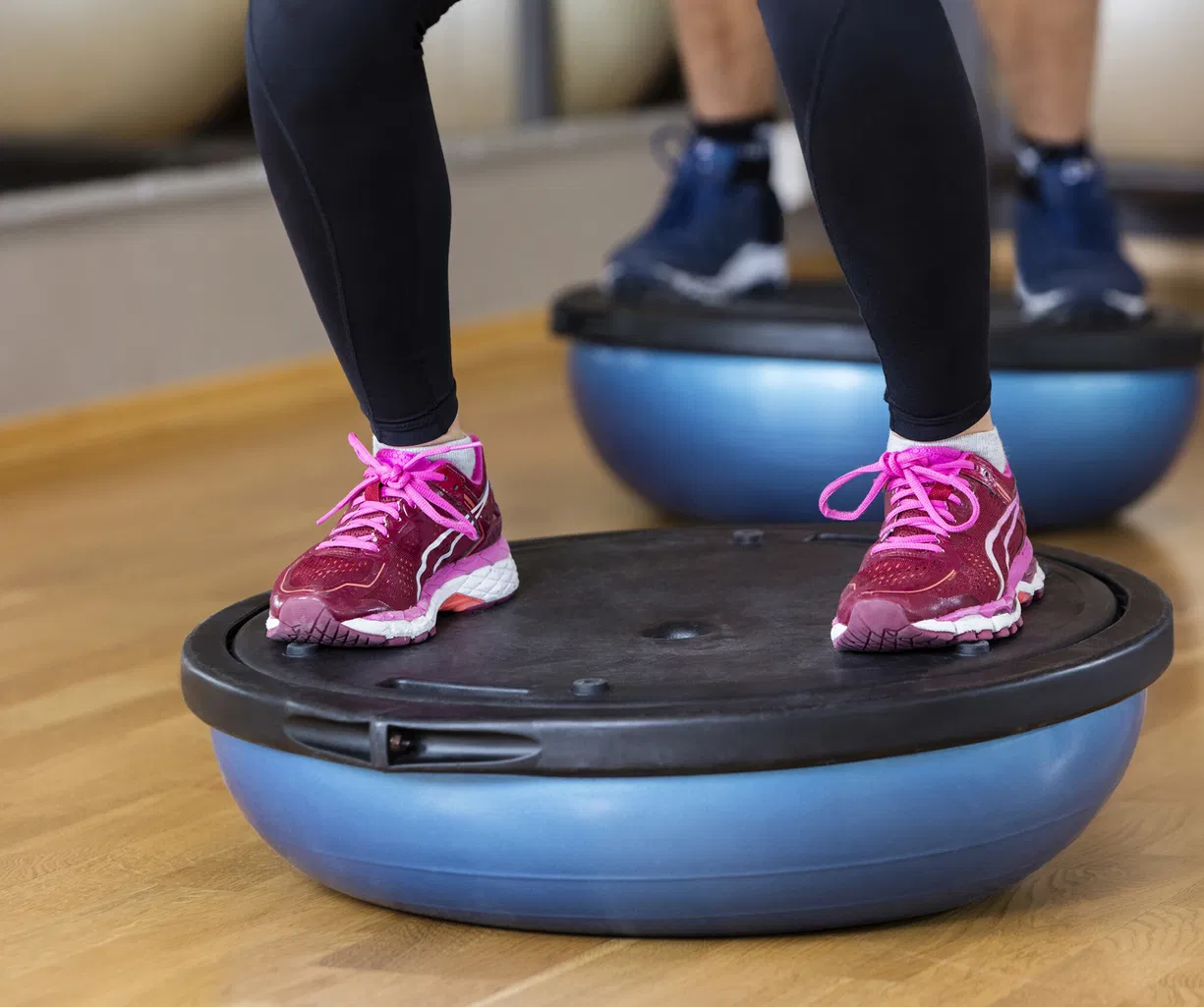 Person in schwarzen Leggings und pinken Sportschuhen steht auf einem Balance-Trainer; im Hintergrund eine weitere Person auf einem ähnlichen Gerät.