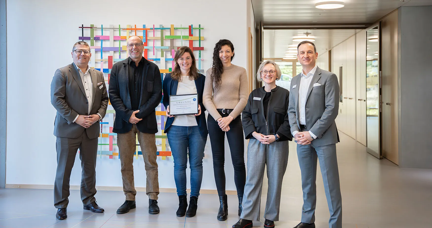 Sechs Personen stehen in einem modernen Innenraum vor einer bunten Wandinstallation. Eine Frau in der Mitte hält ein Zertifikat. Die anderen Personen sind in formeller Kleidung gekleidet und lächeln.