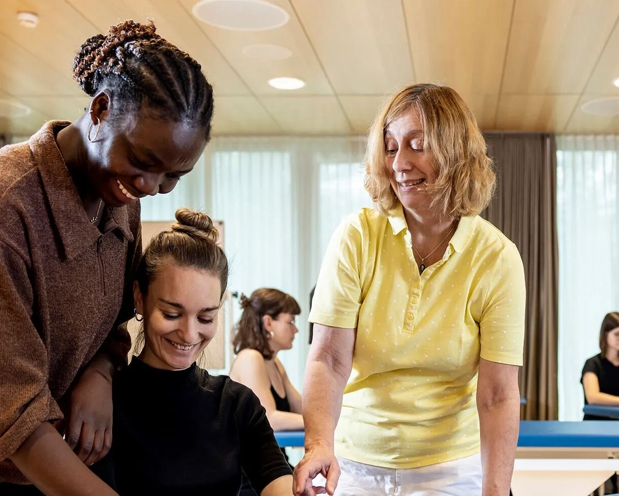Drei Frauen in einem hellen Raum, eine mit lockigem Haar und braunem Oberteil steht hinter einer anderen, die lächelt und ein schwarzes Oberteil trägt. Eine dritte Frau in einem gelben Poloshirt zeigt auf einen Tisch. Im Hintergrund sind weitere Frauen...