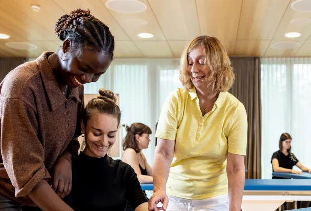 Drei Frauen in einem hellen Raum, eine mit lockigem Haar und braunem Oberteil steht hinter einer anderen, die lächelt und ein schwarzes Oberteil trägt. Eine dritte Frau in einem gelben Poloshirt zeigt auf einen Tisch. Im Hintergrund sind weitere Frauen...