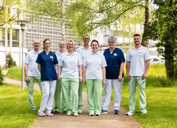Team von acht Mitarbeitenden in einheitlichen Pflegekleidern und grüner bzw. blauer Arbeitskleidung, lächelnd vor einem modernen Gebäude und Grünanlage im Garten.