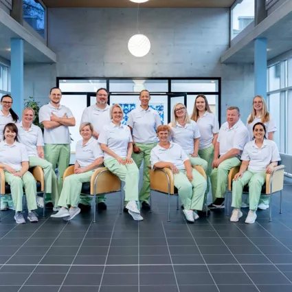 Gruppenfoto von 15 Personen in weißen Oberteilen und hellgrünen Hosen, die in einem modernen Raum mit großen Fenstern und einem hellen Boden stehen und sitzen. Im Hintergrund sind Pflanzen und ein dekoratives Element sichtbar.
