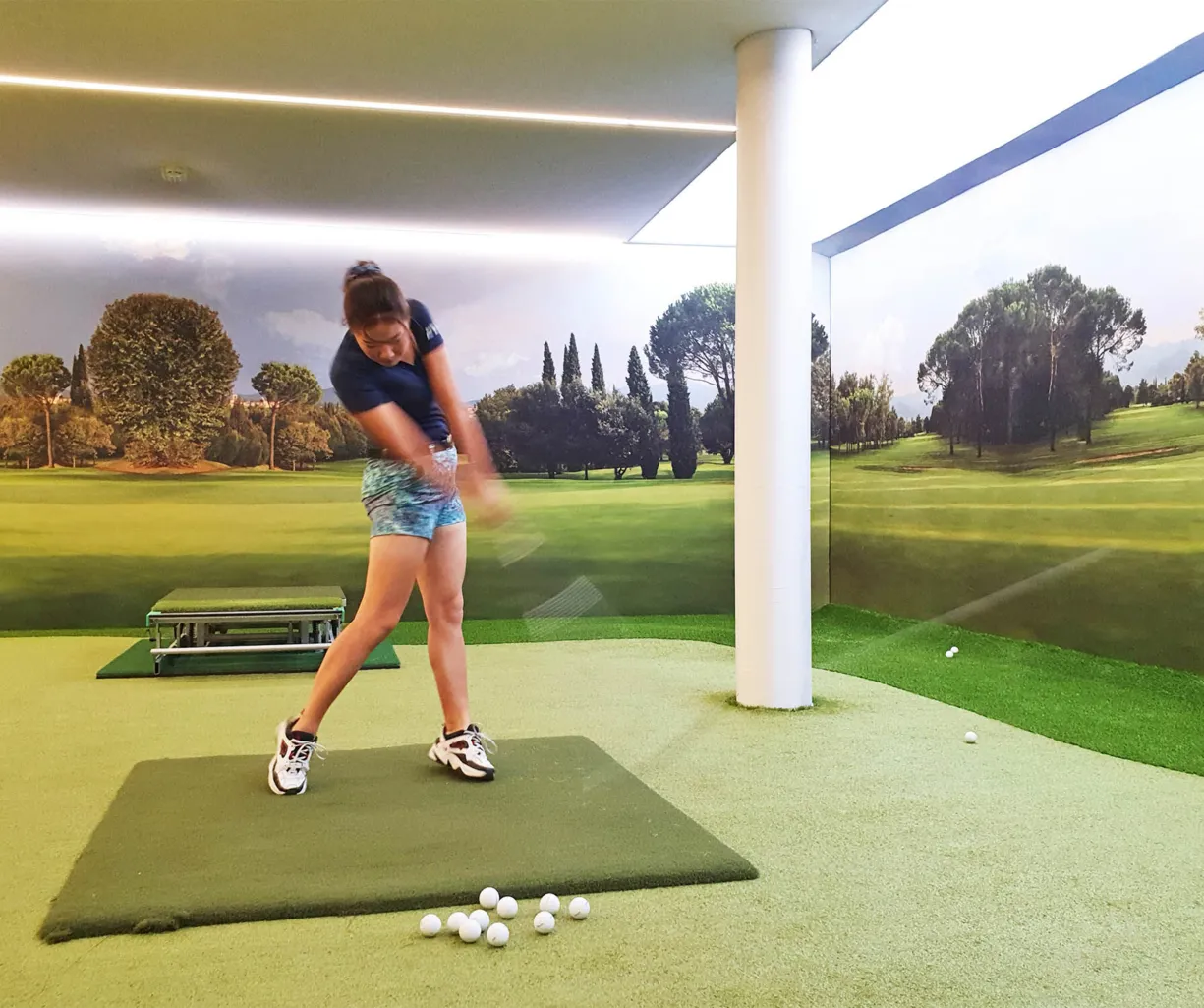 Golfspielerin in einem Indoor-Golfbereich, die einen Schlag ausführt. Im Hintergrund eine Wand mit Landschaftsmotiv, grüne Rasenfläche und mehrere Golfbälle auf dem Boden.