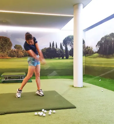 Golfspielerin in einem Indoor-Golfbereich, die einen Schlag ausführt. Im Hintergrund eine Wand mit Landschaftsmotiv, grüne Rasenfläche und mehrere Golfbälle auf dem Boden.