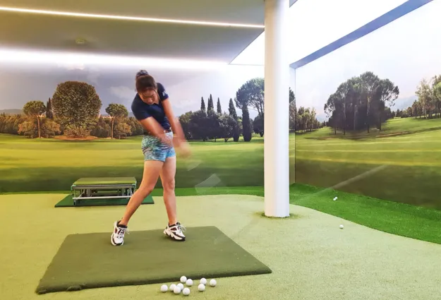 Golfspielerin in einem Indoor-Golfbereich, die einen Schlag ausführt. Im Hintergrund eine Wand mit Landschaftsmotiv, grüne Rasenfläche und mehrere Golfbälle auf dem Boden.