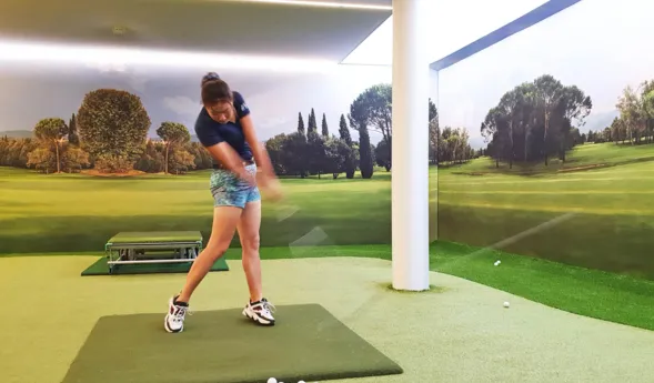 Golfspielerin in einem Indoor-Golfbereich, die einen Schlag ausführt. Im Hintergrund eine Wand mit Landschaftsmotiv, grüne Rasenfläche und mehrere Golfbälle auf dem Boden.