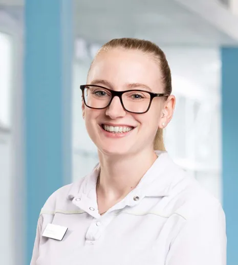 Lächelnde Frau mit Brille, in weißer Arbeitskleidung, vor einem hellen, modernen Hintergrund.