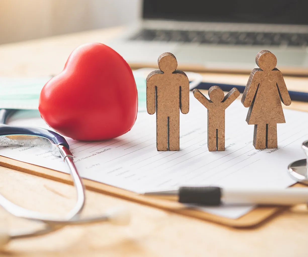 Holzfiguren einer Familie (Mann, Frau, Kind) stehen neben einem roten Herz, einem Stethoskop, einer medizinischen Maske und einem Dokument auf einem Tisch. Im Hintergrund ist ein Laptop sichtbar.