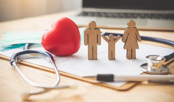 Holzfiguren einer Familie (Mann, Frau, Kind) stehen neben einem roten Herz, einem Stethoskop, einer medizinischen Maske und einem Dokument auf einem Tisch. Im Hintergrund ist ein Laptop sichtbar.